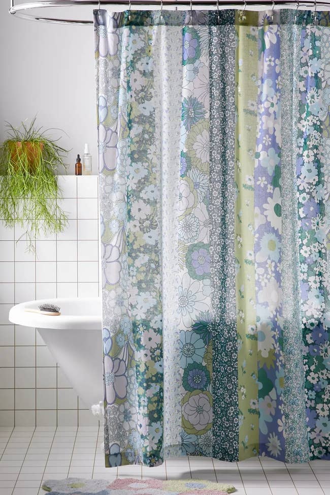 Floral-patterned shower curtain with various flowers and plants in a white-tiled bathroom; bathtub and green hanging plant visible