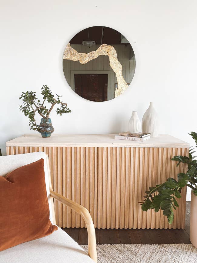 A minimalist room featuring a wooden sideboard with decor items, a round mirror with goldtone accent above, and a chair with an orange cushion