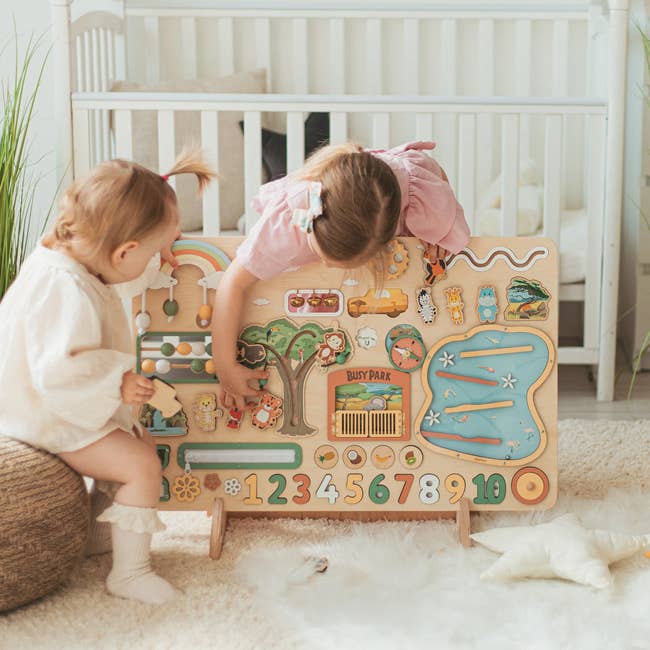 Two young children play with a wooden educational toy board featuring various interactive elements like numbers and puzzles