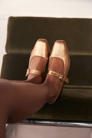 Person wearing gold Mary Jane flats sitting on a dark green velvet chair