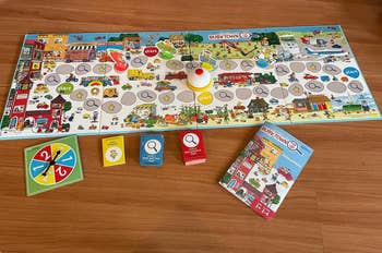 Children's board game set up on a wooden floor, featuring a colorful game board, spinner, cards, and instruction manual. The game theme is busy-town related