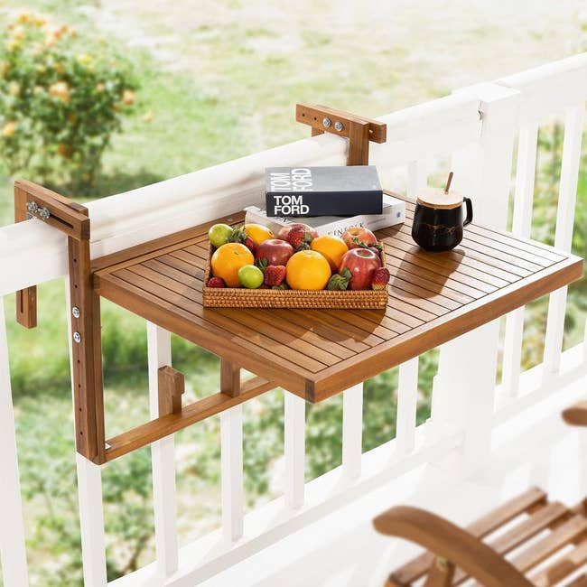 Foldable wooden balcony table with books, a fruit bowl, and a mug, attached to a railing