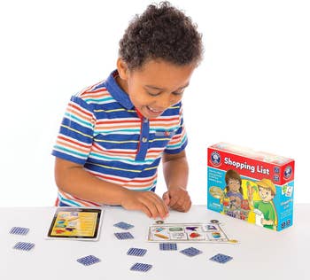 A child plays the Shopping List board game at a table, showing excitement. The game box, featuring illustrations of two animated characters, is also visible