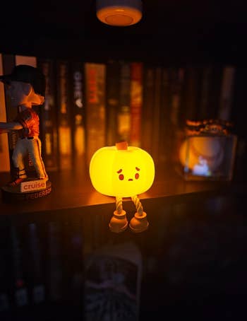 A small pumpkin-shaped night light with a face sits on a shelf surrounded by books, figurines, and a baseball