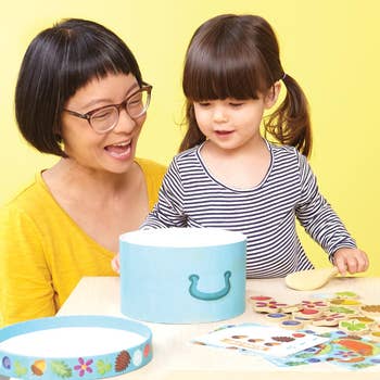 Adult and child playing with a Montessori-inspired toy on a table featuring sorting shapes and a container. They look engaged and happy