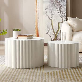 Two cylindrical white ribbed side tables with books and a plant on one and a glass of water on the other in a living room