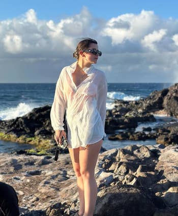 Woman in a sheer beach cover-up, standing on rocky shore with ocean in background. She wears sunglasses and holds a small black item