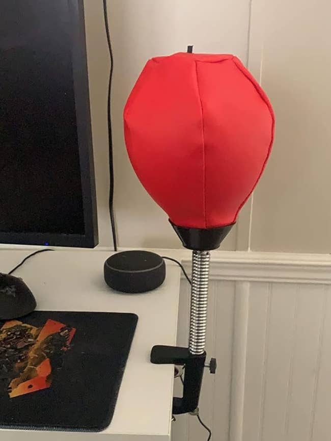 A desktop punching bag with a red balloon-like top, mounted on a table next to a computer monitor and an Amazon Echo device