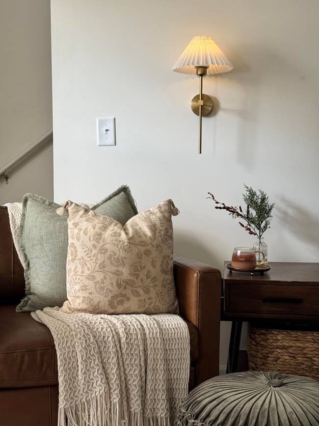 A cozy corner with a leather sofa, decorative pillows, a throw blanket, a wall lamp, side table, and plant