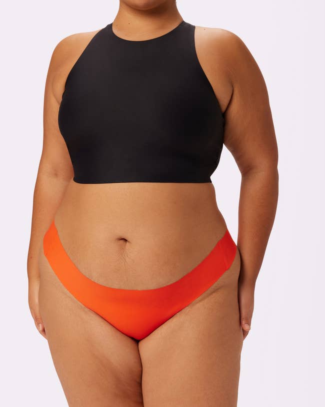 A person wearing a high-neck black crop top and orange briefs, posing for a shopping category article