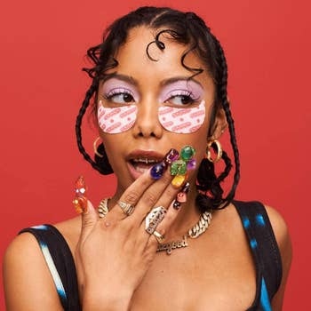 A woman with decorative under-eye patches, colorful nail art, and two braids styled in tight curls, touches her face in a thoughtful pose, against a plain background