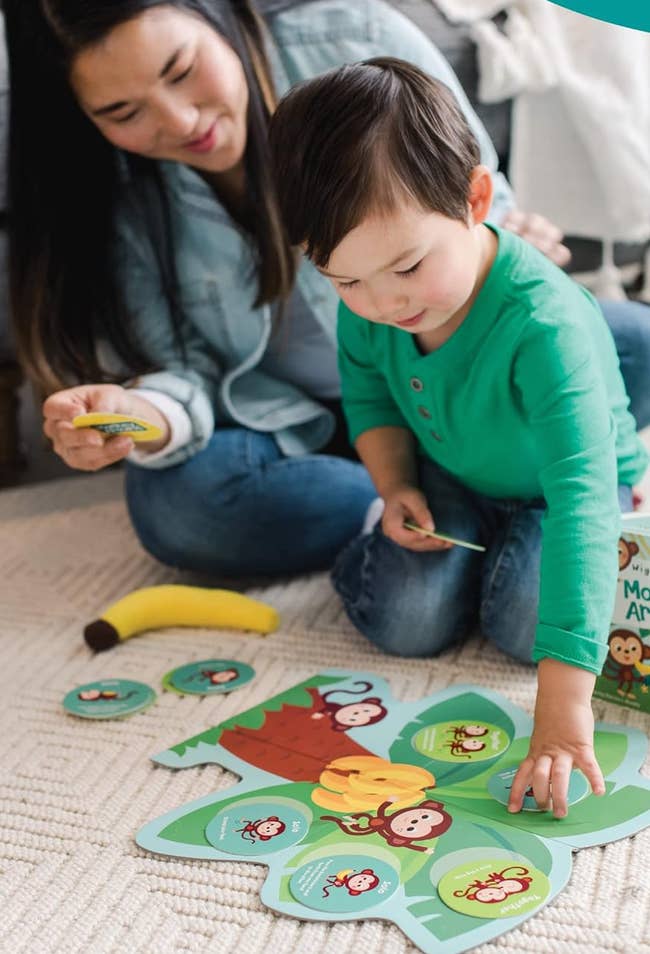 A mother and child play a monkey-themed matching game on the floor. The child is pointing at a card, while the mother watches. A toy banana lies nearby