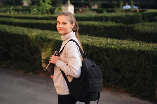 Bezpłatne zdjęcie atrakcyjna uśmiechnięta studentka w przytulnym swetrze z laptopem i plecakiem marzycielsko patrząca w kamerę na zewnątrz