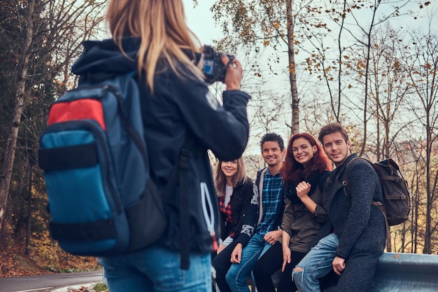 Bezpłatne zdjęcie dziewczyna robi zdjęcie swoim przyjaciołom. grupa młodych przyjaciół siedzi na poręczy w pobliżu drogi. podróże, turystyka, koncepcja przygody.