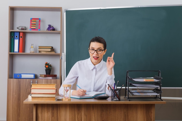 Bezpłatne zdjęcie młoda kobieta nauczyciel w okularach siedzi przy ławce szkolnej przed tablicą w klasie, pisząc w notesie, uśmiechając się wskazując palcem wskazującym w górę, mając świetny pomysł