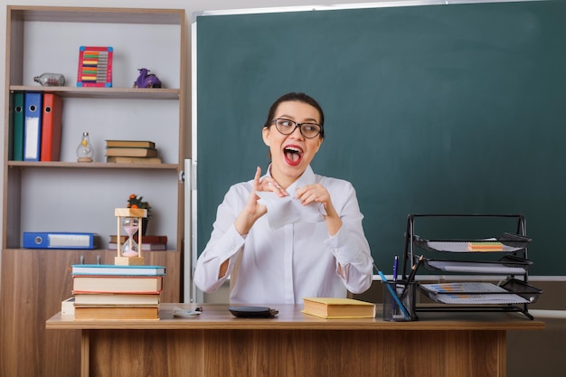 Bezpłatne zdjęcie młoda nauczycielka w okularach rozdziera kartkę papieru i wygląda radośnie siedząc w szkolnej ławce przed tablicą w klasie