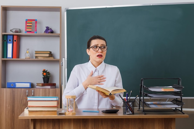 Bezpłatne zdjęcie młoda nauczycielka w okularach siedzi w ławce szkolnej z książką przed tablicą w klasie i wygląda na zmartwioną