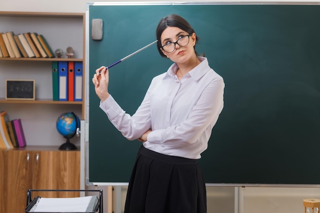 Bezpłatne zdjęcie myśląca patrząc na bok młoda nauczycielka trzymająca wskaźnik na głowie stojąca przed tablicą w klasie