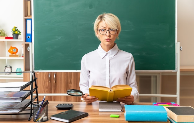 Bezpłatne zdjęcie pod wrażeniem młoda blond nauczycielka w okularach, siedząca przy biurku z szkolnymi narzędziami w klasie, trzymająca otwartą książkę patrząc na kamerę