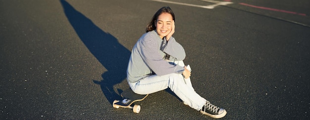 Bezpłatne zdjęcie portret azjatyckiej kobiety siedzącej na deskorolce na jej cruiser longboard za pomocą aplikacji na smartfonie