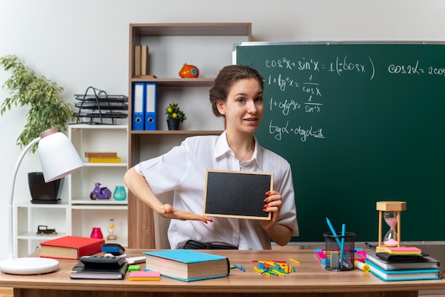 Bezpłatne zdjęcie zadowolona młoda nauczycielka matematyki siedząca przy biurku z przyborami szkolnymi trzymająca i wskazująca na mini tablicę patrzącą na przód w klasie