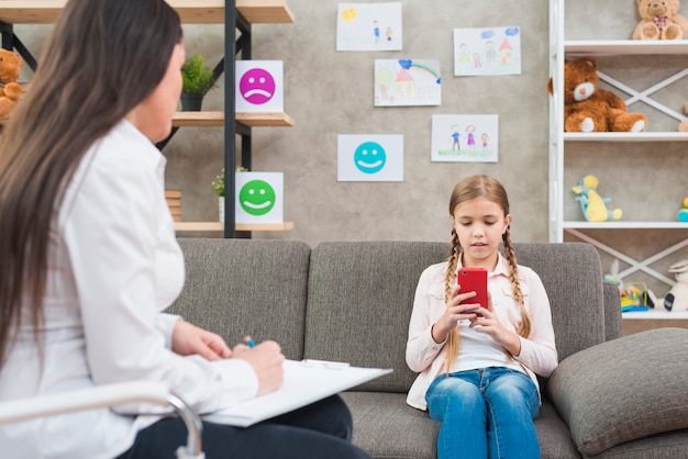 Bezpłatne zdjęcie zakończenie żeńska psychologa writing notatka na schowku przed dziewczyną używa telefon komórkowego