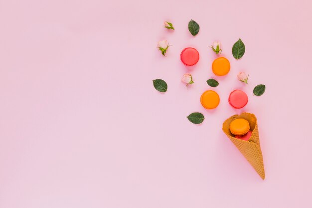 Amaretti colorati; rosa; e foglie disposte sopra il cono della cialda su sfondo rosa