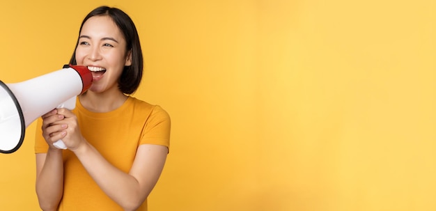 Foto gratuito annuncio donna asiatica felice che grida forte al megafono che recluta protestando con l'altoparlante in mano in piedi su sfondo giallo