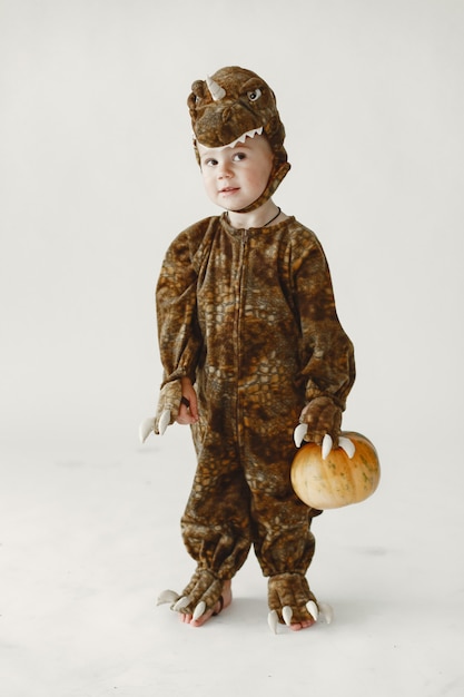 Foto gratuito bambino del ragazzino vestito in costume marrone di un dinosauro che tiene una zucca. il ragazzo ha un cappuccio con la faccia di dinosauro