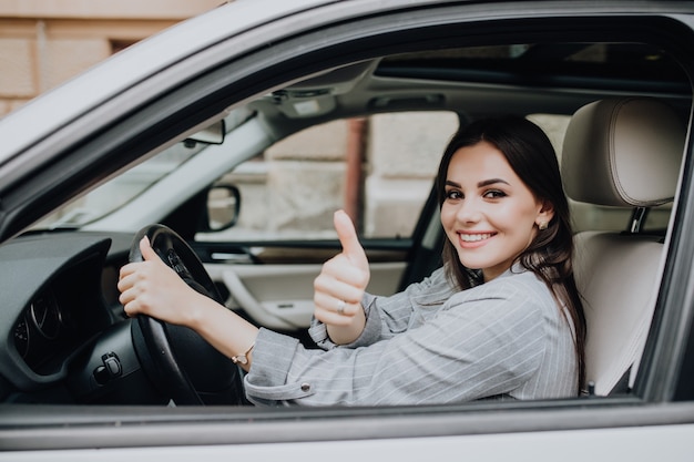 Bella giovane donna latina che guida la sua automobile nuova di zecca e che mostra il suo pollice in su