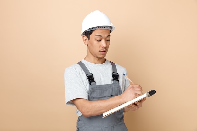 Foto gratuito concentrato giovane operaio edile che indossa il casco di sicurezza e la scrittura uniforme con la matita sul blocco note