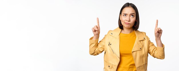 Deluso coreano modello femminile accigliato e smorfie scettico rivolto verso l'alto e guardando sconvolto banner in cima in piedi su sfondo bianco