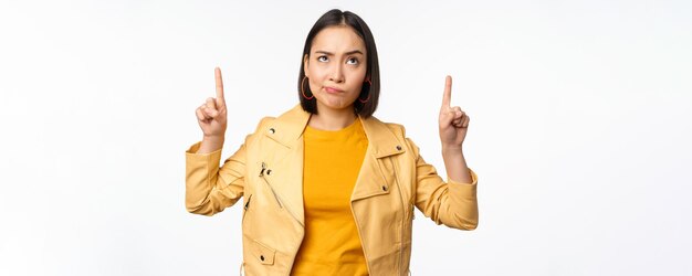 Deluso coreano modello femminile accigliato e smorfie scettico rivolto verso l'alto e guardando sconvolto banner in cima in piedi su sfondo bianco