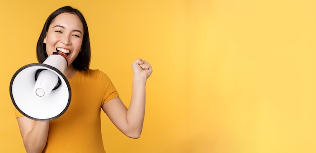 Foto gratuito donna asiatica sorridente in piedi con il megafono che annuncia un prodotto pubblicitario smth in piedi sopra il giallo