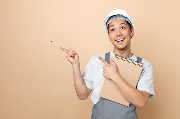 Foto gratuito eccitato giovane operaio edile che indossa il casco di sicurezza e uniforme tenendo il blocco note e matita che osserva in su rivolto verso l'alto all'angolo