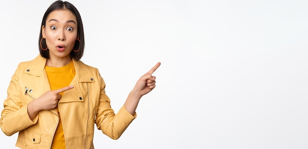 Entusiasta ragazza asiatica che punta il dito verso destra mostrando pubblicità e sorridente invitando al negozio in piedi su sfondo bianco