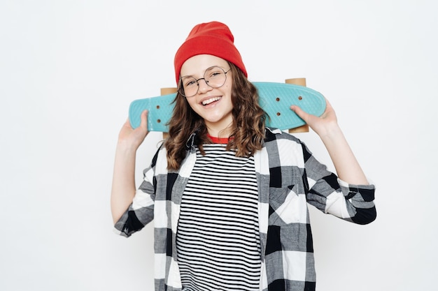 Foto gratuito felice adolescente ragazza alla moda con gli occhiali, berretto rosso, pantaloncini bianchi e camicia a quadri in posa con penny board su bianco