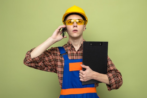 Foto gratuito fiducioso giovane costruttore maschio tenendo appunti indossando l'uniforme con gli occhiali parla al telefono