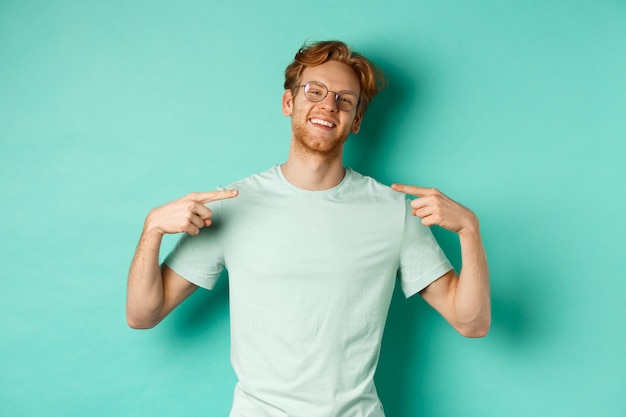 Foto gratuito fiducioso uomo dai capelli rossi con gli occhiali e la maglietta, sorridente con il viso compiaciuto e indicando se stesso, vantandosi in piedi su sfondo turchese.