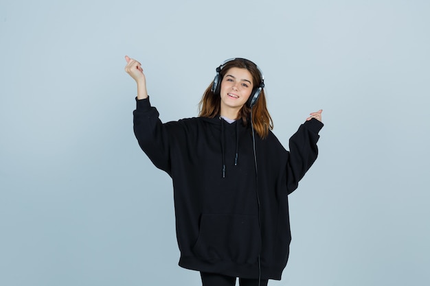 Foto gratuito giovane donna che tiene le mani in alto mentre ascolta la musica con handphones in felpa oversize, pantaloni e sembra energica vista frontale.