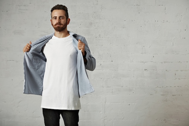 Foto gratuito giovane hipster che si toglie la sua camicia di jeans blu sbiadito che mostra una maglietta di cotone bianco senza etichetta sul muro di mattoni