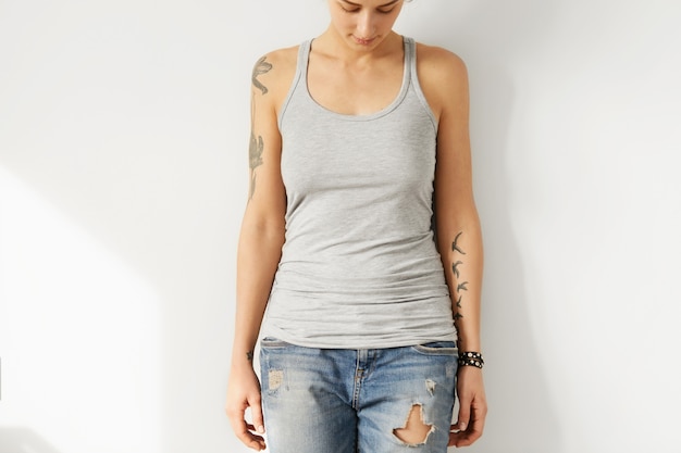 Foto gratuito giovane modello femminile europeo tatuato guardando giù in bianco grigio a-shirt. il ritratto potato della donna dell'adolescente si è vestito in jeans strappati e canottiera sportiva che posano all'interno. gioventù e moda