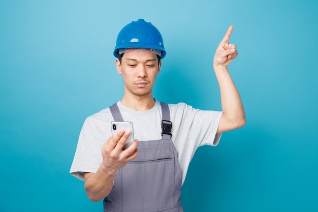 Foto gratuito grave giovane operaio edile che indossa il casco di sicurezza e tenuta uniforme e guardando il telefono cellulare rivolto verso l'alto