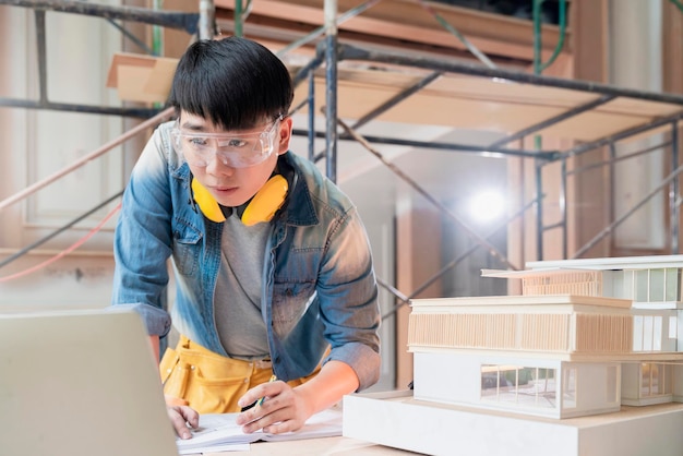 Foto gratuito ingegnere asiatico maschio con uniforme che lavora con laptop e modello di casa modello analisi risultato di costruzione con tecnologia applicativa in cantiere casa ristrutturare e miglioramento sfondo