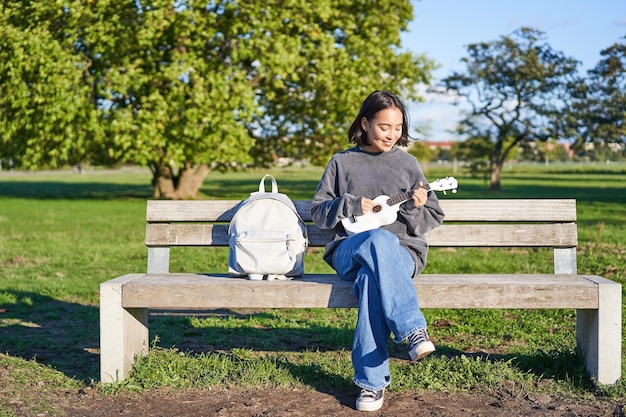 Foto gratuito la ragazza spensierata si siede sulla panchina nel parco con l'ukulele suona e canta all'aperto in una giornata di sole felice