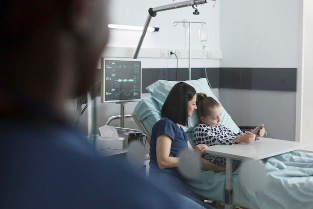 Foto gratuito madre seduta accanto al letto del paziente nella stanza della clinica pediatrica mentre una bambina carina che gioca sullo smartphone. giovane paziente gioioso che si gode l'intrattenimento del telefono cellulare mentre è seduto nel reparto di recupero della clinica.