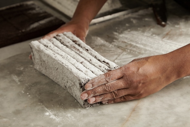 Mani dello chef uomo nero tiene barrette di cioccolato appena sfornate in polvere di zucchero prima del confezionamento, primo piano in pasticceria artigianale professionale