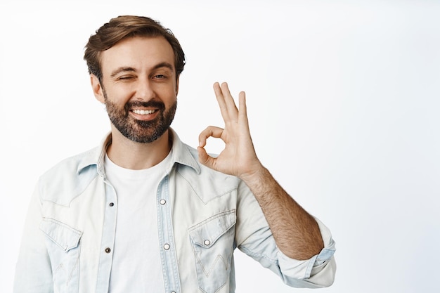 Foto gratuito molto bene sorridente bell'uomo mostra segno ok e strizza l'occhio approva smth dare il permesso raccomanda buon prodotto sfondo bianco