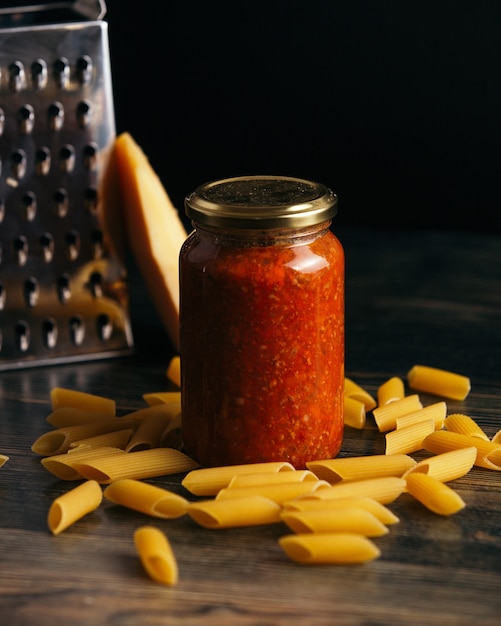 Foto gratuito penne e un barattolo di salsa sul tavolo con formaggio e una grattugia sullo sfondo