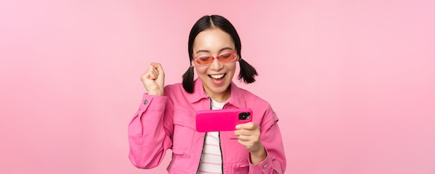 Foto gratuito ragazza coreana sorridente felice che vince sul telefono cellulare guardando lo schermo orizzontale dello smartphone e gioendo per raggiungere l'obiettivo celebrando in piedi su sfondo rosa